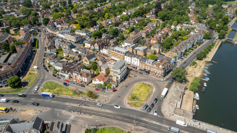 Aerial view of a coastal town showing a mixture of residential and commercial buildings, with a main road running through the centre and lined with parked cars. Several white vans and moving trucks are parked or positioned along the street, indicating recent or ongoing home relocation activity. The image captures a small roundabout near the waterfront, where some vehicles are turning or waiting. To the right, a harbour area with boats docked along the water’s edge is visible, with a concrete promenade and pathways adjacent to the shoreline. Surrounding the town are lush green trees and residential houses with tiled roofs, set amongst greenery and streets. The scene is well-lit with natural daylight, highlighting the urban layout and the movement of vehicles involved in packing, loading, or furniture transport as part of a professional removals process, with Man and Van Hampton Hill providing relocation services nearby.
