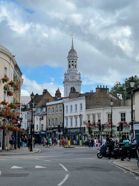 A busy street scene in Hampton Hill showing a mix of historic and modern buildings, with storefronts on the ground floor and residential windows above. Several pedestrians are walking along the pavement, some transiting across the road, while others browse shop entrances or wait at bus stops. A black scooter is parked on the right side, and a group of people are gathered at a bus stop near the corner. The street is lined with black lampposts and hanging flower baskets. In the background, there is a large, historic clock tower with a spire extending into the cloudy blue sky, providing a focal point that emphasizes the area's traditional architecture. The environment appears lively but orderly, with vehicles including a van visible near the street curb. This scene reflects the typical activity during a home relocation or furniture transport operation in Hampton Hill, with some individuals potentially involved in packing or moving activities supported by Man and Van Hampton Hill. The lighting suggests daylight hours, with natural light illuminating the scene clearly. 
