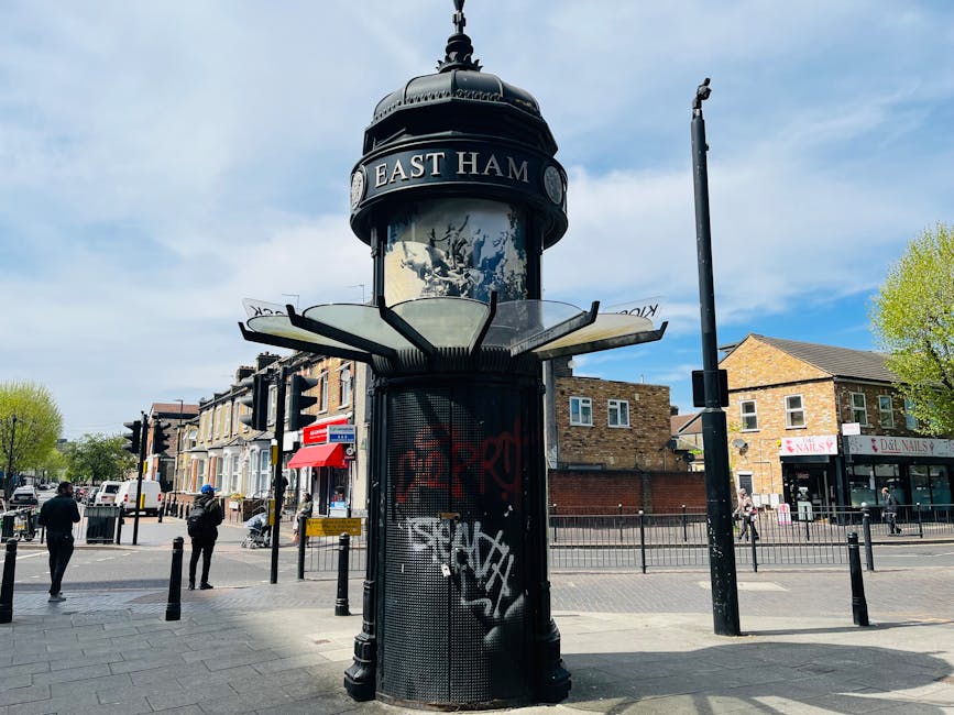 A black, vintage-style cylindrical public notice board with a domed top and a decorative finial, situated at a street corner in East Ham. The notice board features a glass display area at the top, currently obscured by a wrap-around sign reading 'EAST HAM,' and a perforated metal panel below with graffiti. The area surrounding the notice board includes some pedestrians walking on the pavement, bollards, a traffic light, and a small shopfront with signage. In the background, there are residential buildings with brick facades and commercial premises, along with a partly cloudy sky and green trees. The scene captures an urban environment suitable for home relocation planning or furniture transport considerations, with an emphasis on the street's community atmosphere.