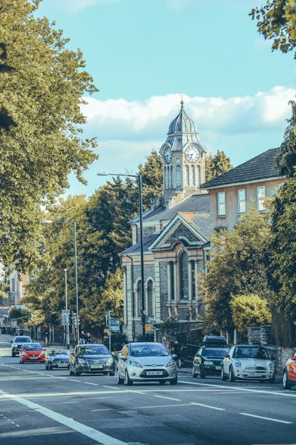 A busy street scene in Hampton Hill showing a mix of historic and modern buildings, with storefronts on the ground floor and residential windows above. Several pedestrians are walking along the pavement, some transiting across the road, while others browse shop entrances or wait at bus stops. A black scooter is parked on the right side, and a group of people are gathered at a bus stop near the corner. The street is lined with black lampposts and hanging flower baskets. In the background, there is a large, historic clock tower with a spire extending into the cloudy blue sky, providing a focal point that emphasizes the area's traditional architecture. The environment appears lively but orderly, with vehicles including a van visible near the street curb. This scene reflects the typical activity during a home relocation or furniture transport operation in Hampton Hill, with some individuals potentially involved in packing or moving activities supported by Man and Van Hampton Hill. The lighting suggests daylight hours, with natural light illuminating the scene clearly. 