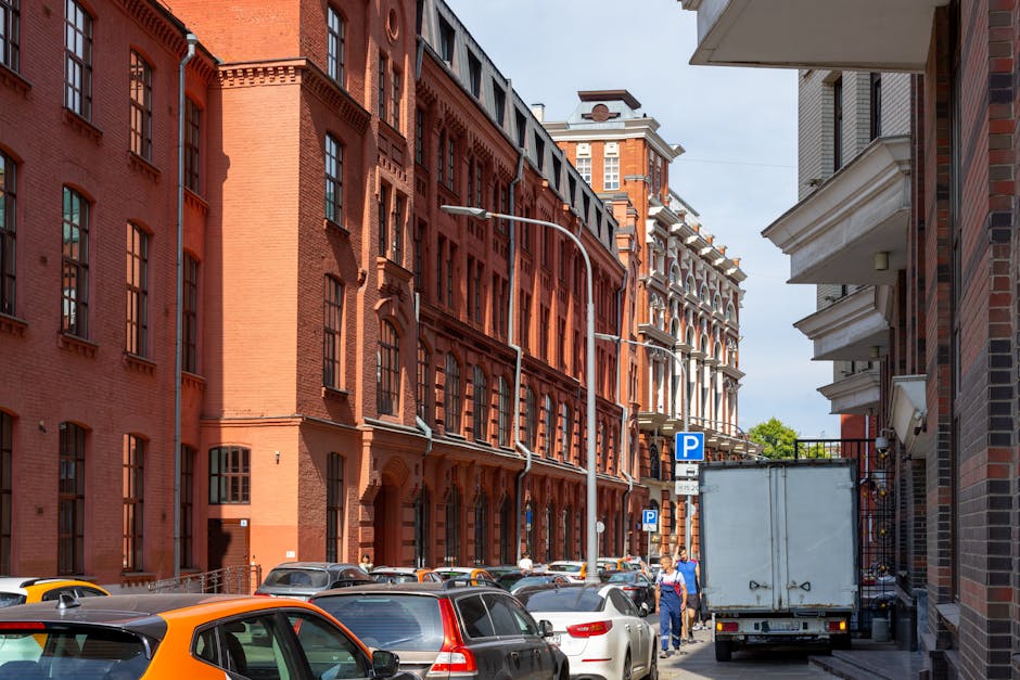 A narrow urban street scene showing parked cars lining both sides of the road, including a black, orange, and white vehicle, with a white delivery truck partially visible on the right side, adjacent to a historical red-brick building with large multi-pane windows. Street lighting fixtures are mounted onto the buildings, and a blue parking sign is visible near the truck, indicating parking restrictions. The scene takes place outdoors during daytime, with clear weather and shadows cast on the buildings. The surroundings suggest a dense residential or commercial area, typical for house removals or furniture transport in Hampton Hill. Man and Van Hampton Hill may utilize such street views for planning or navigating during home relocation, loading, and packing logistics within tight city streets congested with parked vehicles, highlighting potential parking challenges for removals.