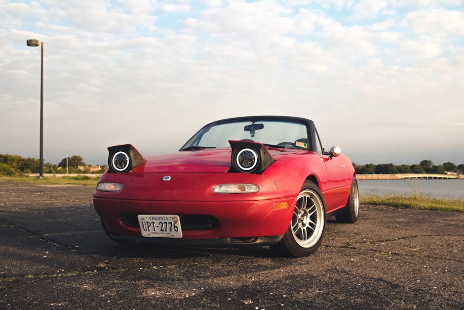 A red convertible sports car with its headlights removed and placed on the front hood, parked on an asphalt surface near a body of water under a partly cloudy sky. The car has an open top, silver alloy wheels, and a Virginia license plate reading UPT-2776. In the background, there is a lamppost on the left side, some greenery, and a distant bridge over the water. The scene appears to be outdoors during daylight, possibly during a home relocation or vehicle transport process. The image is captured to showcase the vehicle's exterior details, focusing on the car's positioning in a parking or loading area, relevant to house removals or moving logistics as managed by Man and Van Hampton Hill.