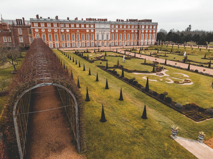 An aerial view of the formal gardens and landscaped grounds of Hampton Court Palace, with neatly trimmed lawns, symmetrical flowerbeds, and small decorative pathways. In the foreground, there is a gravel pathway leading through a garden archway covered with bare, woody vines, indicating winter season. Tall, narrow, dark green conical shrubs line the edges of the lawns, and several classical statues and ornamental features are visible within the garden. The background features the historic red-brick palace building with white window frames and detailed architectural features, set against a cloudy sky. The scene captures the well-maintained outdoor environment, suitable for home relocation or furniture transport services, with open spaces and pathways that could accommodate moving equipment, staff, or delivery activities. Man and Van Hampton Hill occasionally mentions their removals services that facilitate house moves, including planning and logistics for access to historic sites like Hampton Court Palace.