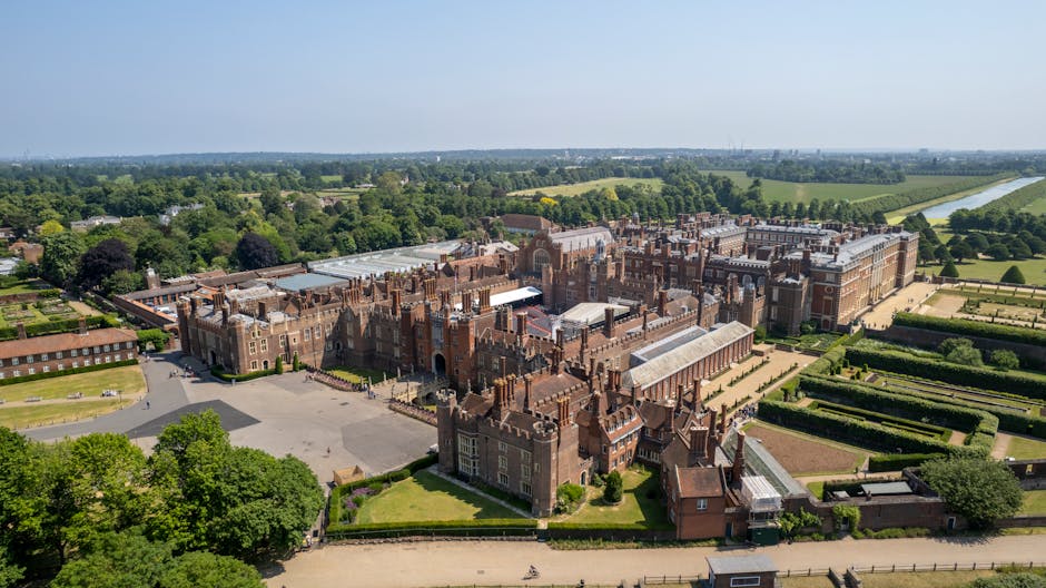 An aerial view of the formal gardens and landscaped grounds of Hampton Court Palace, with neatly trimmed lawns, symmetrical flowerbeds, and small decorative pathways. In the foreground, there is a gravel pathway leading through a garden archway covered with bare, woody vines, indicating winter season. Tall, narrow, dark green conical shrubs line the edges of the lawns, and several classical statues and ornamental features are visible within the garden. The background features the historic red-brick palace building with white window frames and detailed architectural features, set against a cloudy sky. The scene captures the well-maintained outdoor environment, suitable for home relocation or furniture transport services, with open spaces and pathways that could accommodate moving equipment, staff, or delivery activities. Man and Van Hampton Hill occasionally mentions their removals services that facilitate house moves, including planning and logistics for access to historic sites like Hampton Court Palace.