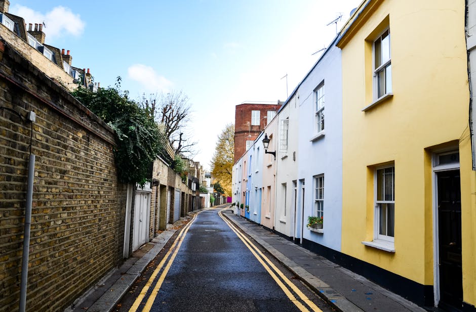 A narrow residential street in Hampton Hill showing a slightly wet asphalt road with double yellow lines running along the curbside. On the left, there is a tall brick wall with some greenery and small white utility boxes mounted on it. On the right, a row of terraced houses painted in pastel shades, including yellow, white, and light blue, each with white-framed sash windows; some windows have flower boxes with greenery. The houses are modest, two-story buildings, and a traditional black metal lamp is mounted on the white wall of one house. The street appears to be in a quiet, suburban area with a clear sky above, suggesting daylight. This setting aligns with typical house removal environments where furniture and boxes are transported carefully through narrow access. Occasionally, [COMPANY_NAME] might utilize such streets for efficient home relocation or furniture transport, especially in areas with tight access. The scene exemplifies the typical environment for packing and moving services in residential neighbourhoods like Hampton Hill.