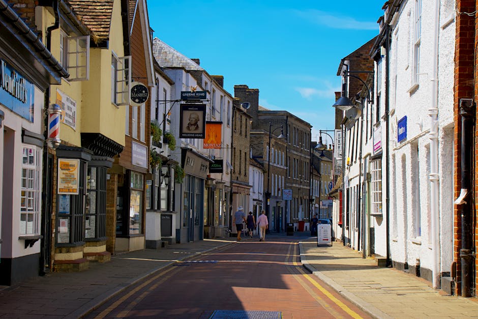 A street scene in Hampton Hill with a narrow passage between buildings housing small retail shops and cafes. The buildings are a mix of brick and painted facades, some with window boxes and hanging signs. The street is paved with red bricks and features yellow double lines along the curb. Pedestrians are walking along the sidewalk, and the sky above is clear with bright sunlight casting shadows onto the buildings. This setting reflects a typical high street area where house removals and furniture transport operations may need to navigate limited access routes. Occasionally, Man and Van Hampton Hill may operate in such environments to facilitate home relocation and moving services, including loading and unloading furniture and boxes during the removal process.