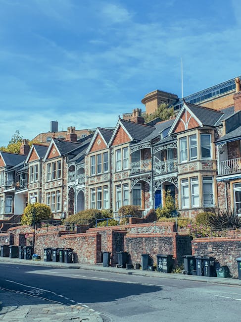 A residential street in Hampton Hill with rows of terraced houses on both sides, each featuring small front gardens with greenery and trees. The street surface is paved with asphalt, and parked cars including various models and colours line both sides of the road, leaving a narrow central lane for traffic. In the background, larger green trees are visible, creating a canopy over parts of the street under a cloudy sky. Utility wires run overhead across the scene, and some houses display rooflines with chimneys. This image captures the typical environment where home relocation and furniture transport services by Man and Van Hampton Hill, such as removals, are often coordinated, especially when navigating limited access or parking restrictions during packing and moving operations.