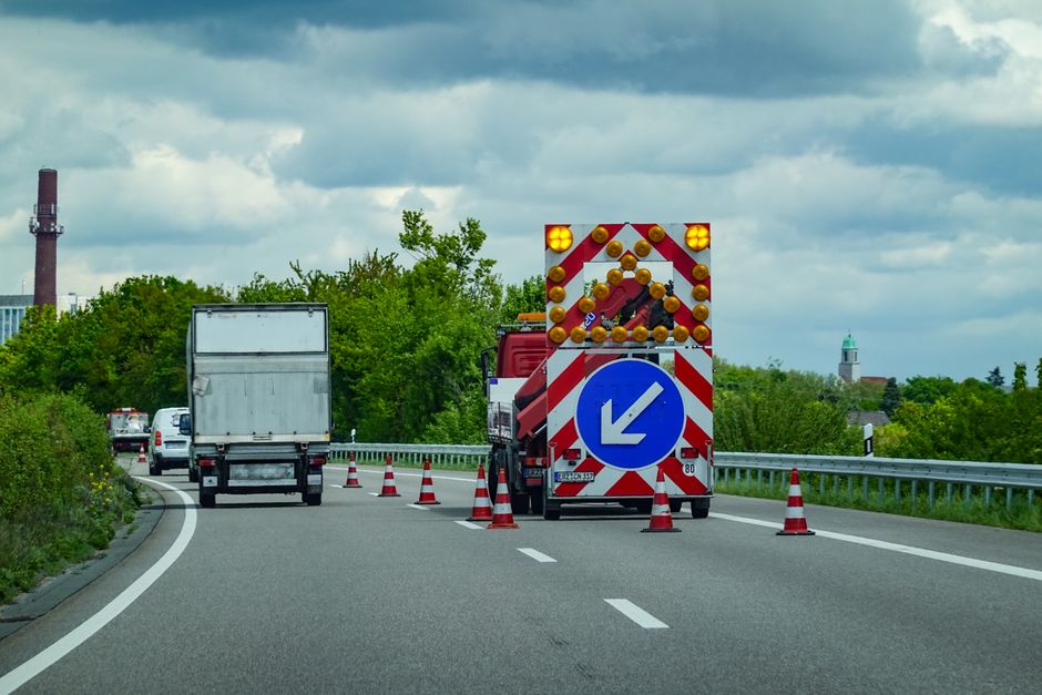 A roadwork vehicle with flashing amber lights and a large blue arrow sign directing traffic to the left is positioned on a dual carriageway, surrounded by orange and white traffic cones. Several vehic