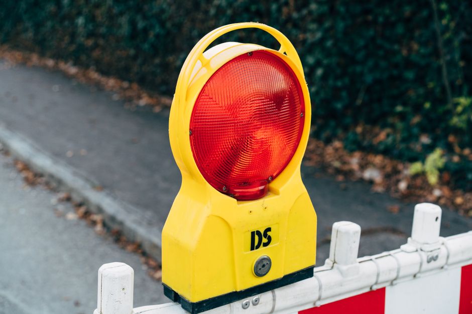A roadwork vehicle with flashing amber lights and a large blue arrow sign directing traffic to the left is positioned on a dual carriageway, surrounded by orange and white traffic cones. Several vehic