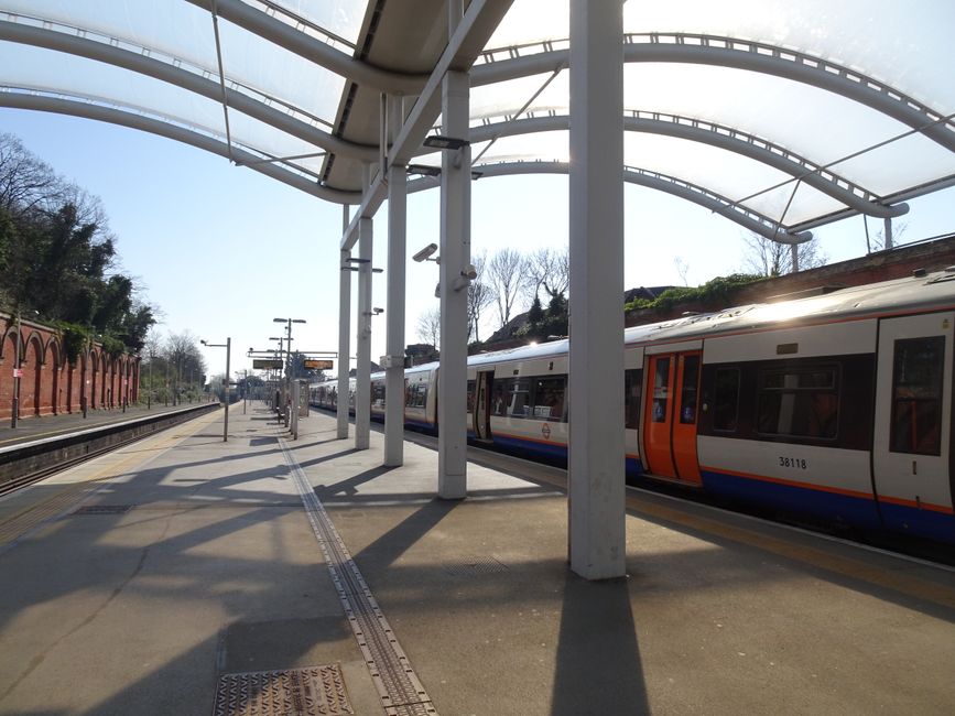 View of St Margarets train station platform on a clear day with bright sunlight casting shadows from the canopy structure supported by white columns. A modern train with a white body, orange and blue