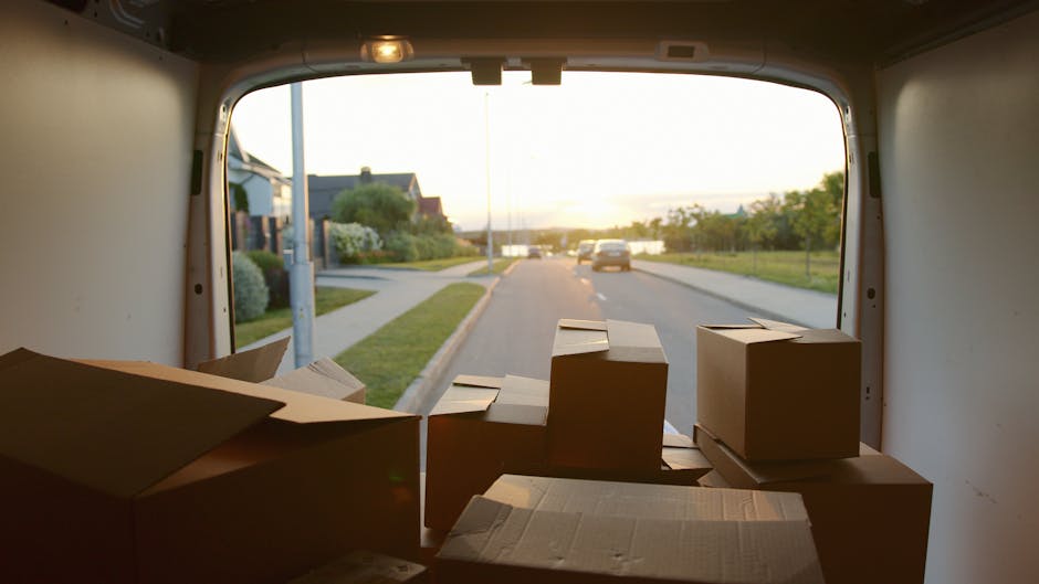 View from inside a delivery van showing a partially loaded interior with several brown cardboard boxes of various sizes arranged on the floor and against the walls, ready for home relocation or furniture transport. Outside the open rear doors, a residential street with pavement, modern houses, greenery, and parked cars is visible during sunset, indicating the final stage of packing and load-in for a house move. The van is situated on a quiet suburban road, with natural light illuminating the scene, emphasizing the process of packing and moving logistics. This setup suggests careful wrapping and organization typical of professional removals services like those offered by Man and Van Hampton Hill, supporting efficient and secure furniture transport and packing services for Hampton Hill residents.
