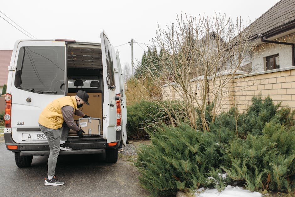 View from inside a delivery van showing a partially loaded interior with several brown cardboard boxes of various sizes arranged on the floor and against the walls, ready for home relocation or furniture transport. Outside the open rear doors, a residential street with pavement, modern houses, greenery, and parked cars is visible during sunset, indicating the final stage of packing and load-in for a house move. The van is situated on a quiet suburban road, with natural light illuminating the scene, emphasizing the process of packing and moving logistics. This setup suggests careful wrapping and organization typical of professional removals services like those offered by Man and Van Hampton Hill, supporting efficient and secure furniture transport and packing services for Hampton Hill residents.